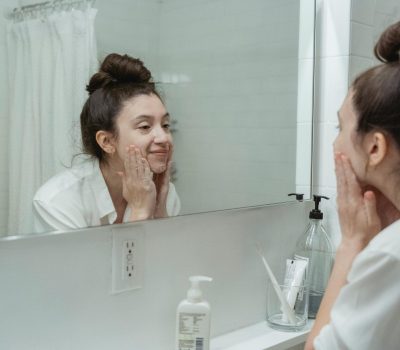 Woman performing skincare routine in a bathroom, applying face cream while looking into a mirror.